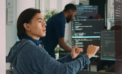 Two software developers working in an office, with one person focused on code displayed across multiple monitors while another collaborates in the background, suggesting active development and teamwork.