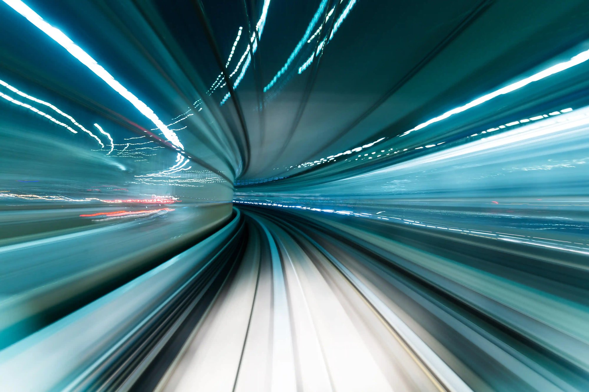 Long-exposure photo of a futuristic high-speed train or metro tunnel with teal and white light trails, creating a dynamic motion blur and sense of rapid transit through a curved underground corridor.