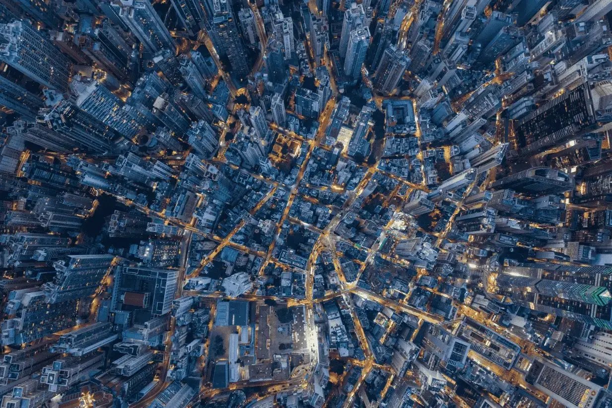 Bird’s-eye view of a dense city at night, with tall buildings packed closely together and glowing streetlights forming a web of illuminated roads between the skyscrapers.