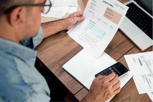 A person sitting at a wooden desk reviews a printed utility bill while using a calculator on a smartphone; papers, a notebook, and a laptop are spread out on the desk, suggesting they are checking or calculating expenses.