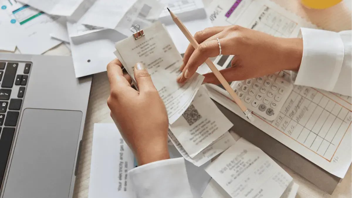 Person reviewing paper receipts and expenses at a desk with calculator, laptop, and financial documents for bookkeeping or budgeting.