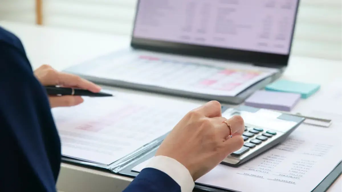 Accountant calculating financial data using a calculator while reviewing documents beside a laptop.