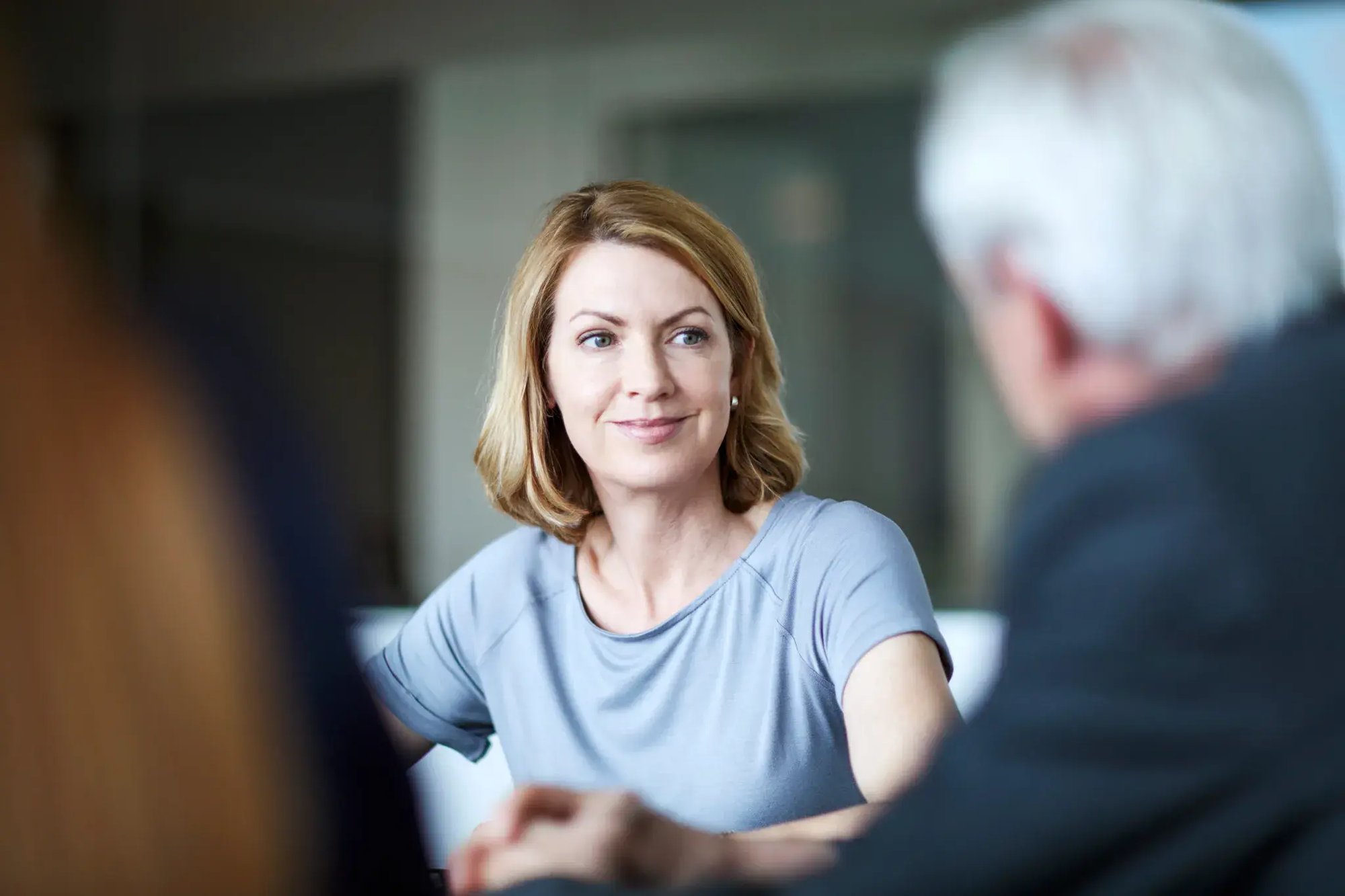 A woman with shoulder-length blonde hair sits at a table, looking attentively at an older man during a conversation in a softly lit indoor setting, suggesting a calm, engaged discussion.