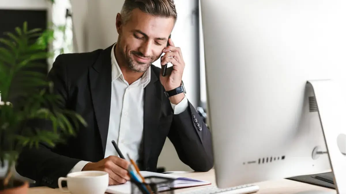 Business professional talking on the phone while taking notes at his office desk with a computer.
