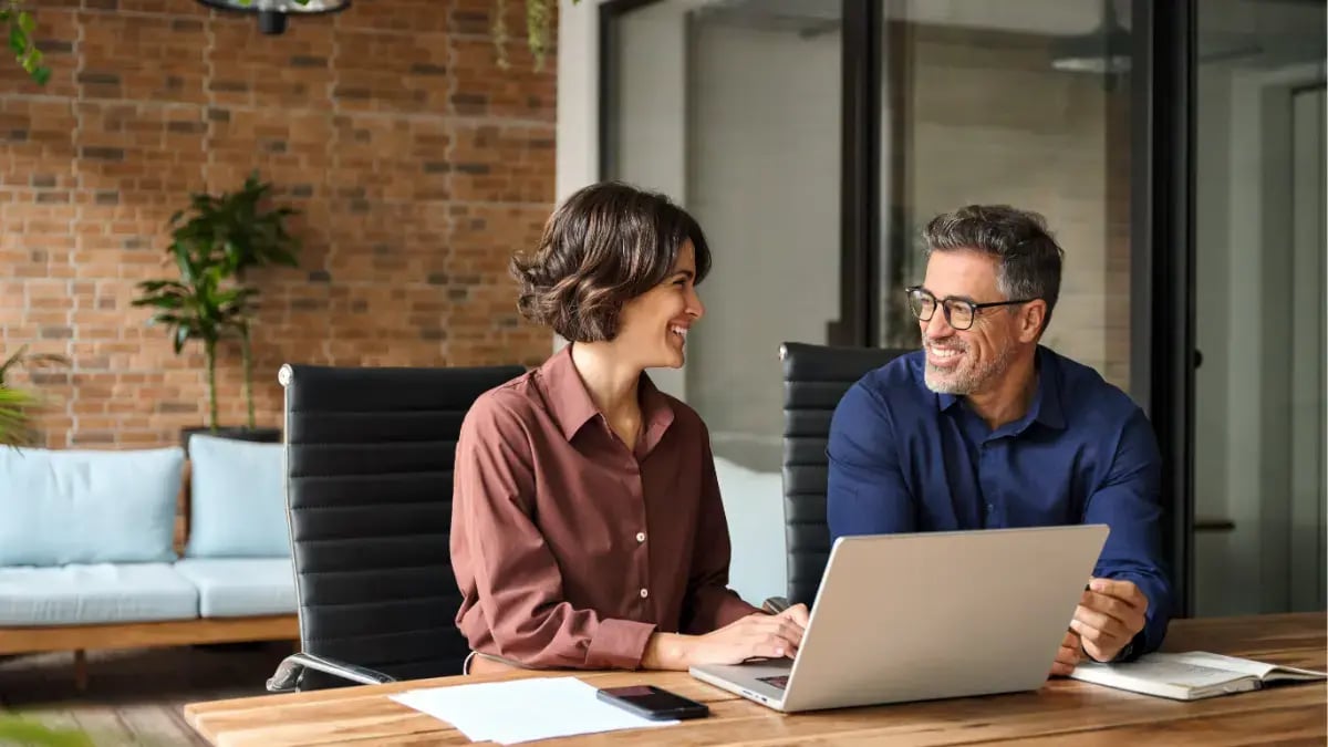 Two colleagues discussing work and collaborating on a laptop in a modern office environment.
