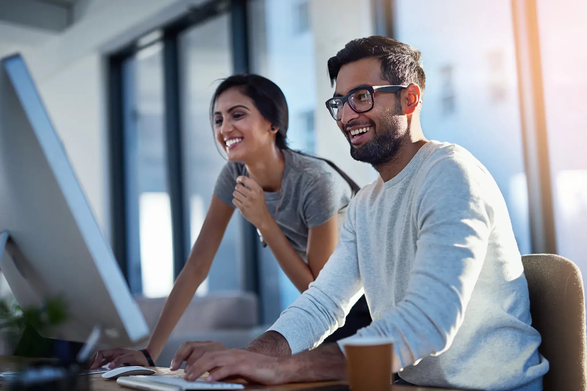 Two colleagues smiling and collaborating at a desktop computer in a modern office environment.