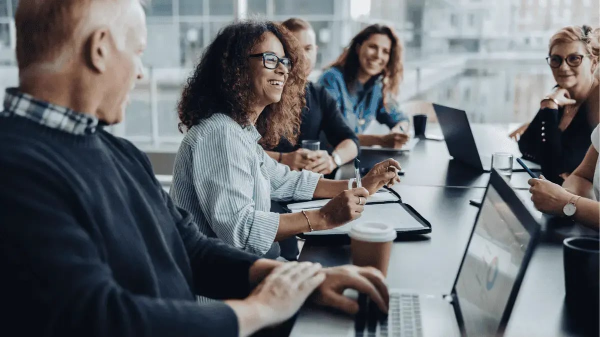 Team of diverse professionals collaborating in a modern office meeting, discussing ideas around a conference table with laptops and notebooks.